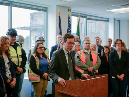 Washington Governor Bob Ferguson speaks at a podium to announce the cost estimate while a group of people stand behind him indoors. Several people wear hard hats and safety vests, and flags are visible in the background.
