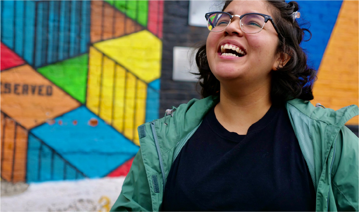 Female wearing glasses and a green shirt standing in front of a brightly colored mural