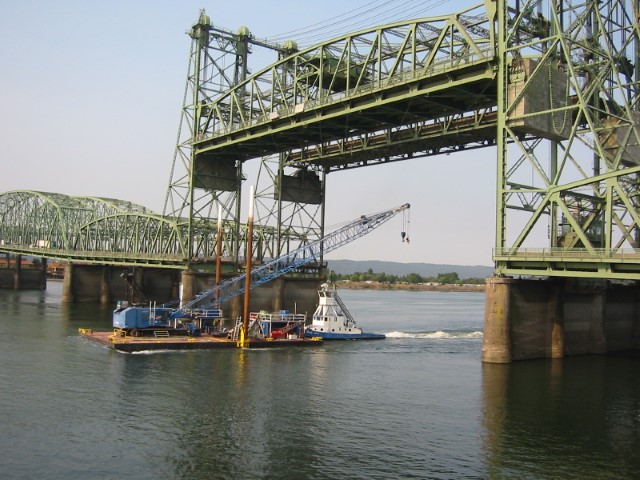 The Interstate Bridge with a ship passing under while a truck and car are driving across the bridge.